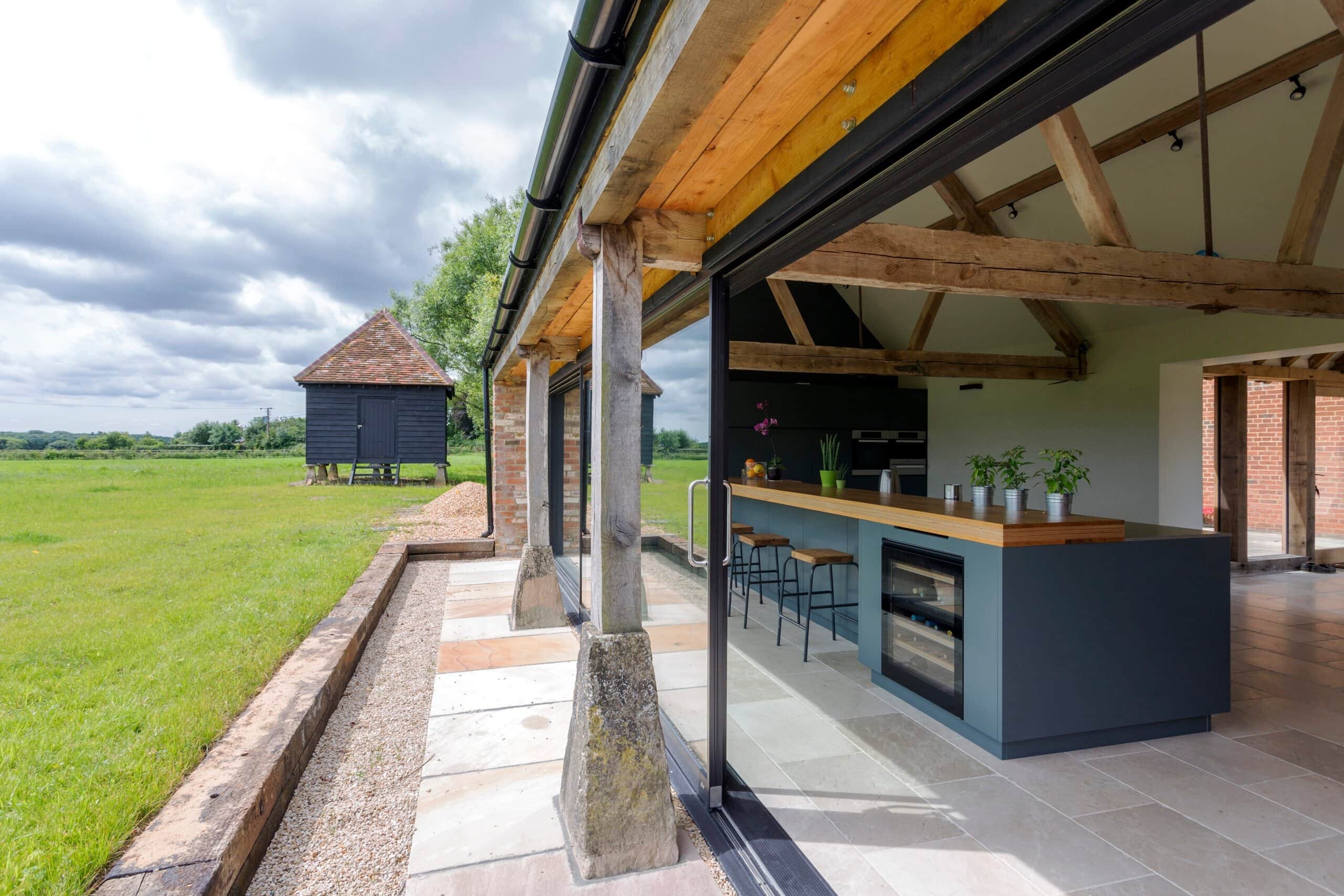 A modern barn conversion featuring an open-plan kitchen with a large teal island, exposed timber beams, and massive sliding glass doors that open to a green countryside view.