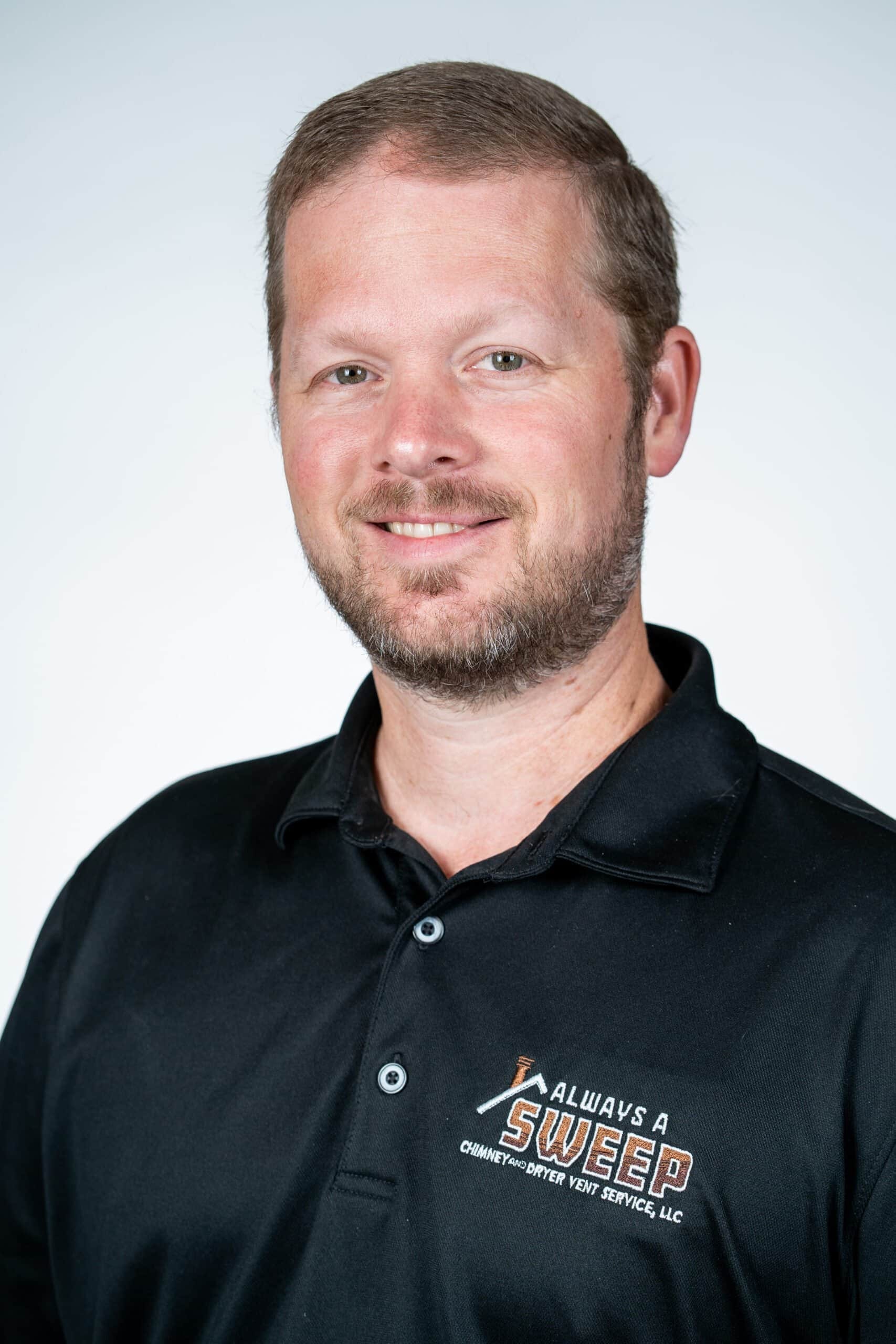 A professional headshot of a smiling man with a beard wearing a black polo shirt featuring the 