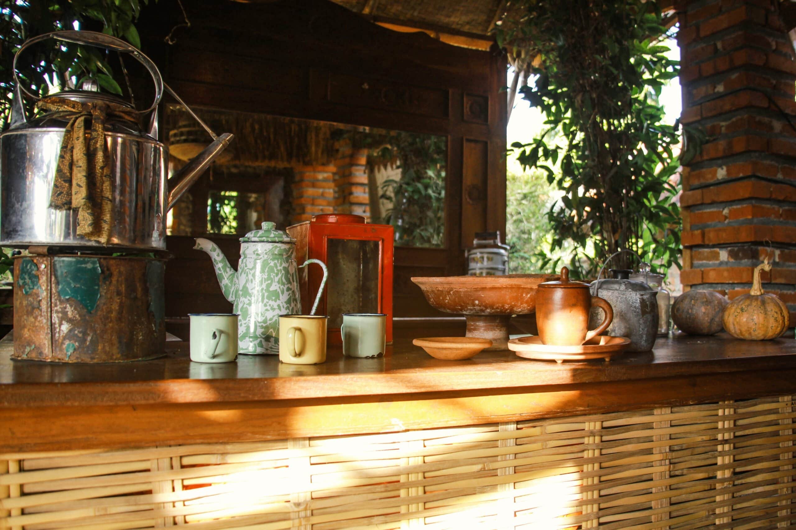 A wooden outdoor counter displaying vintage enamel teapots, colorful mugs, traditional clay pottery, and pumpkins against a bamboo and brick background.