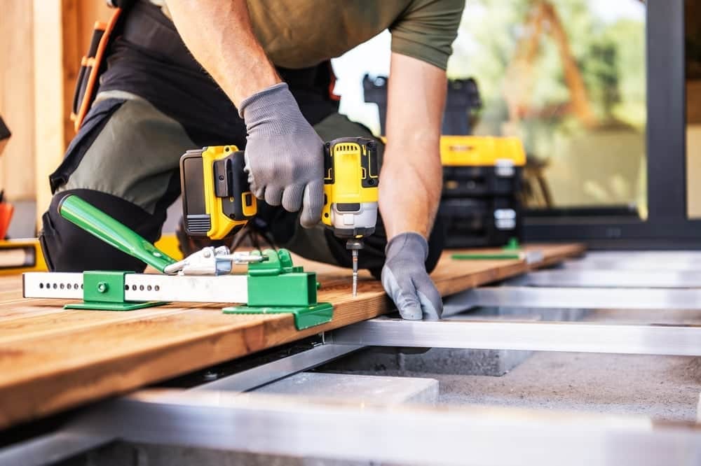 A contractor in work gear kneeling on a wooden deck, using a yellow cordless impact driver and a green board-straightening tool to secure deck planks.
