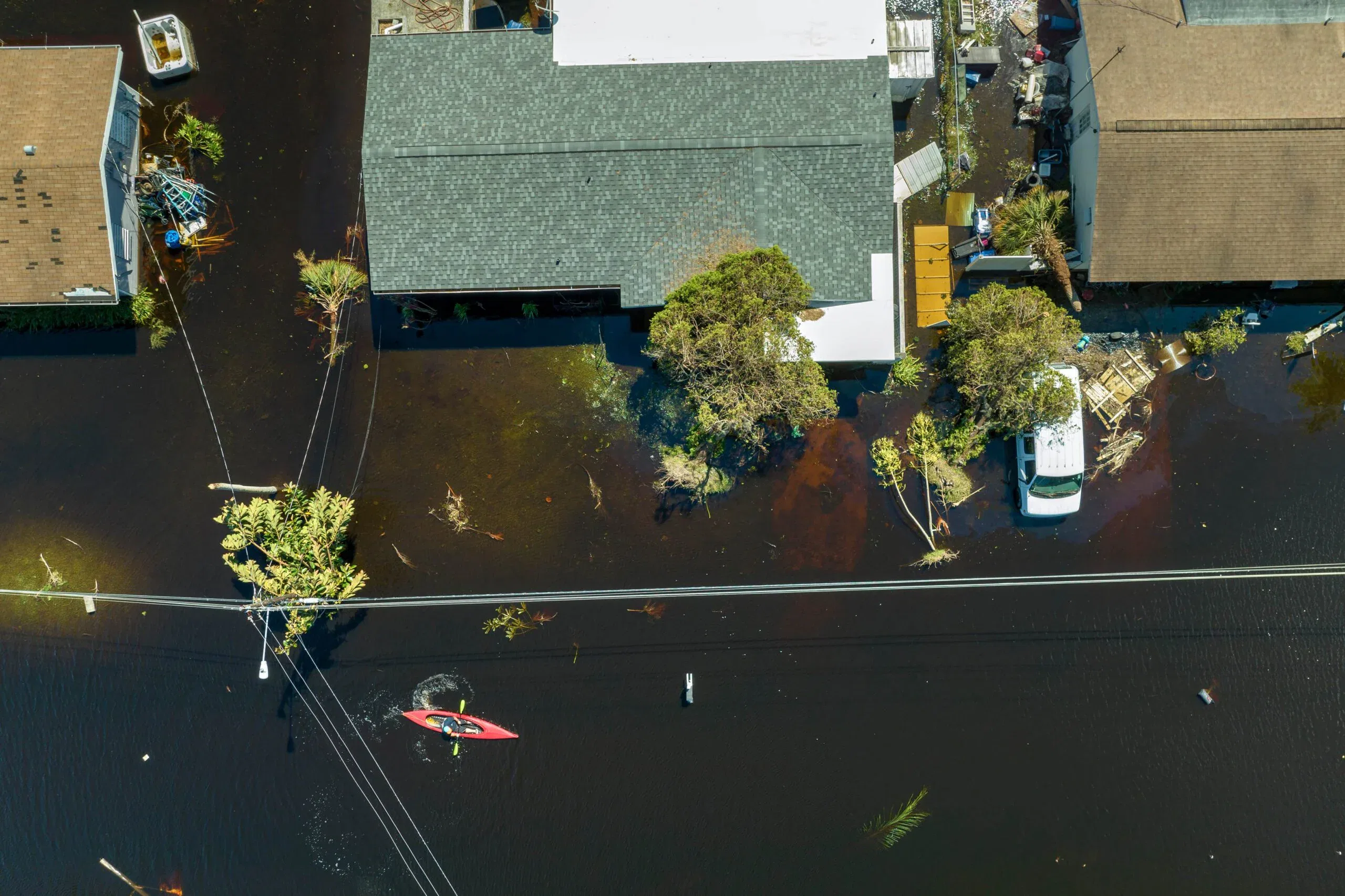 An aerial drone view of a flooded residential street with dark water surrounding houses and a person navigating a red kayak past submerged power lines.