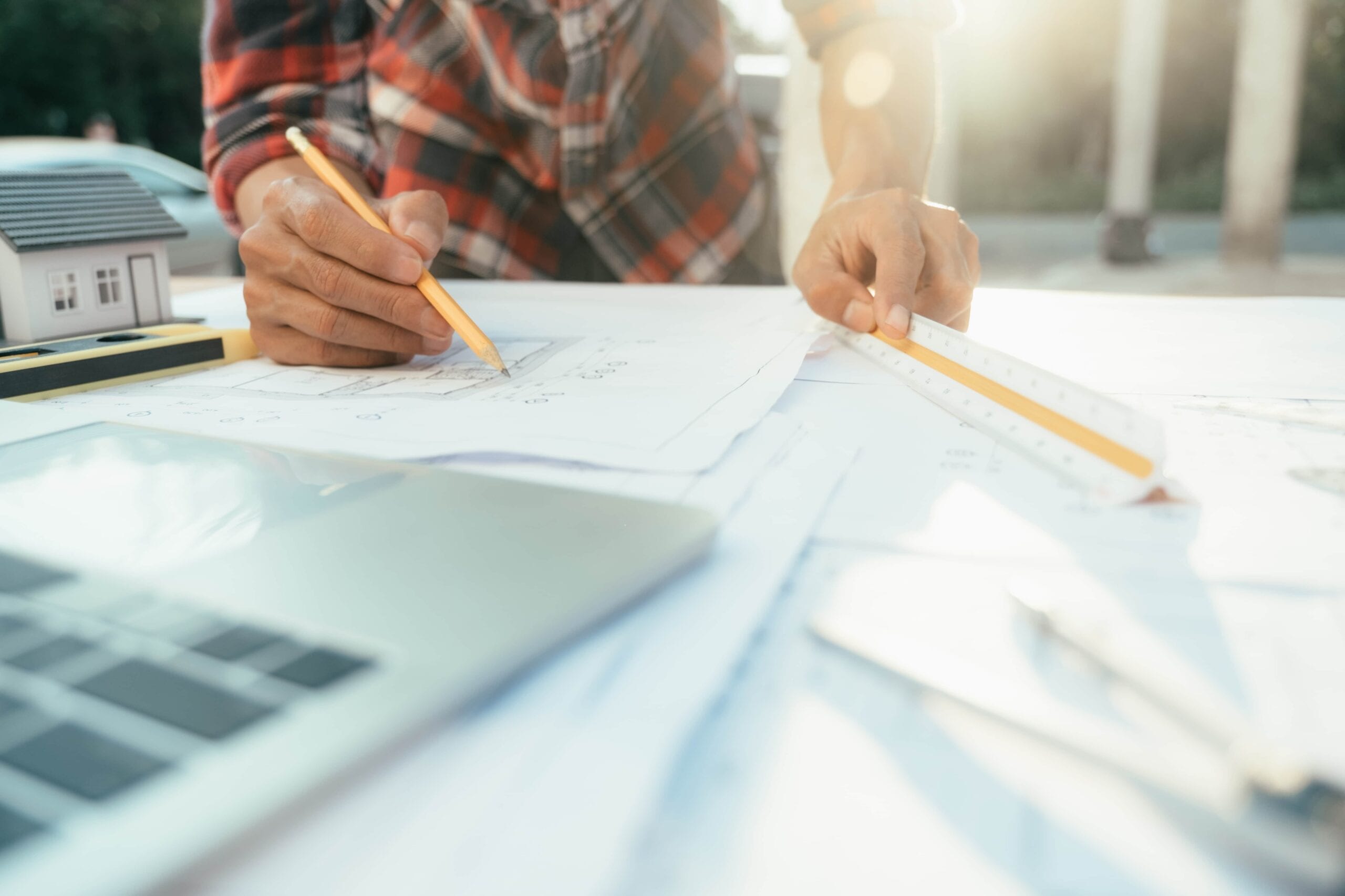 Close-up of an architect and engineer working on residential house blueprints with a pencil, scale ruler, and model home on a desk during sunset.