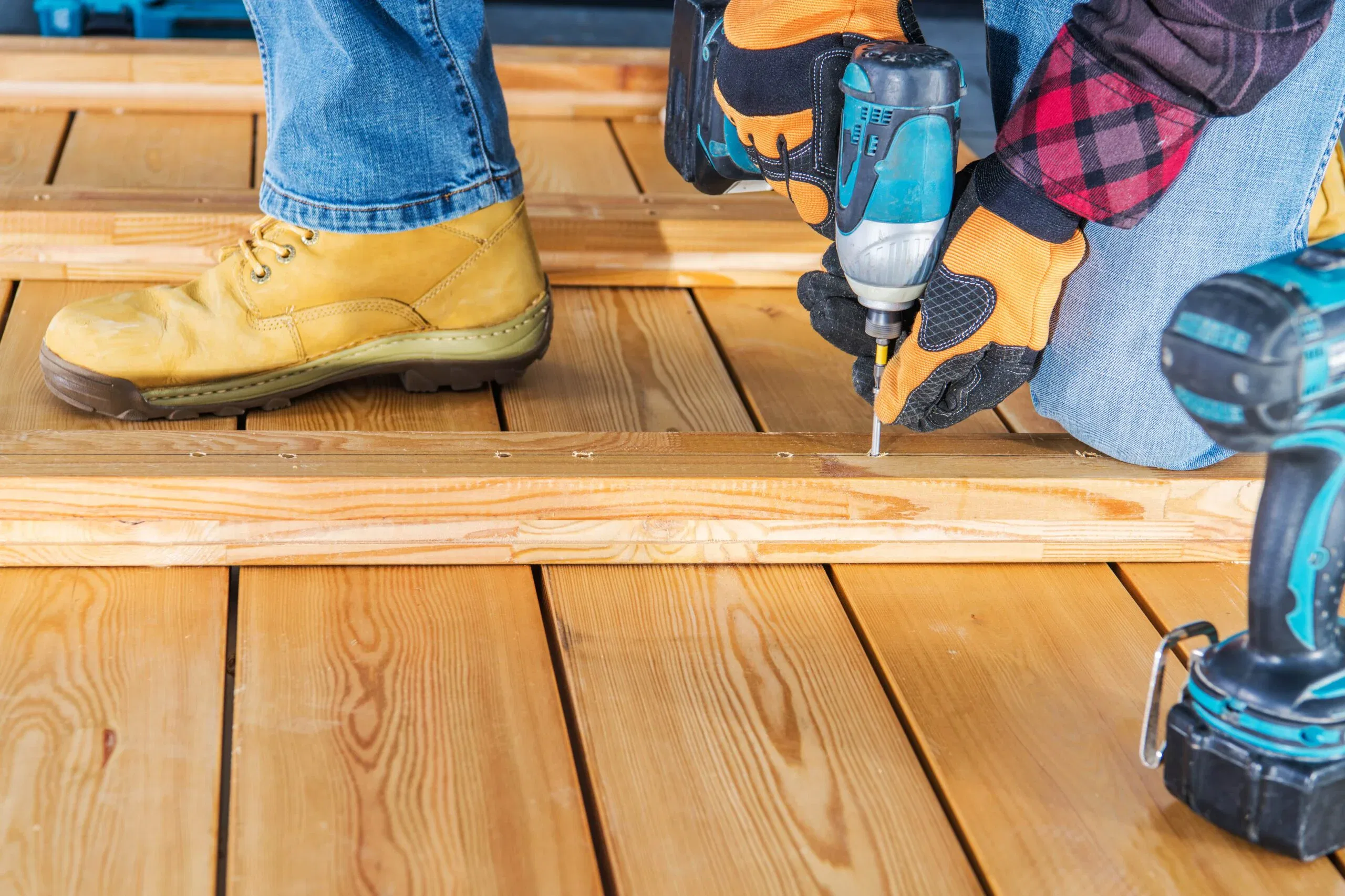 A person wearing work gloves and safety boots using a cordless impact driver to screw a wooden plank onto a deck frame.