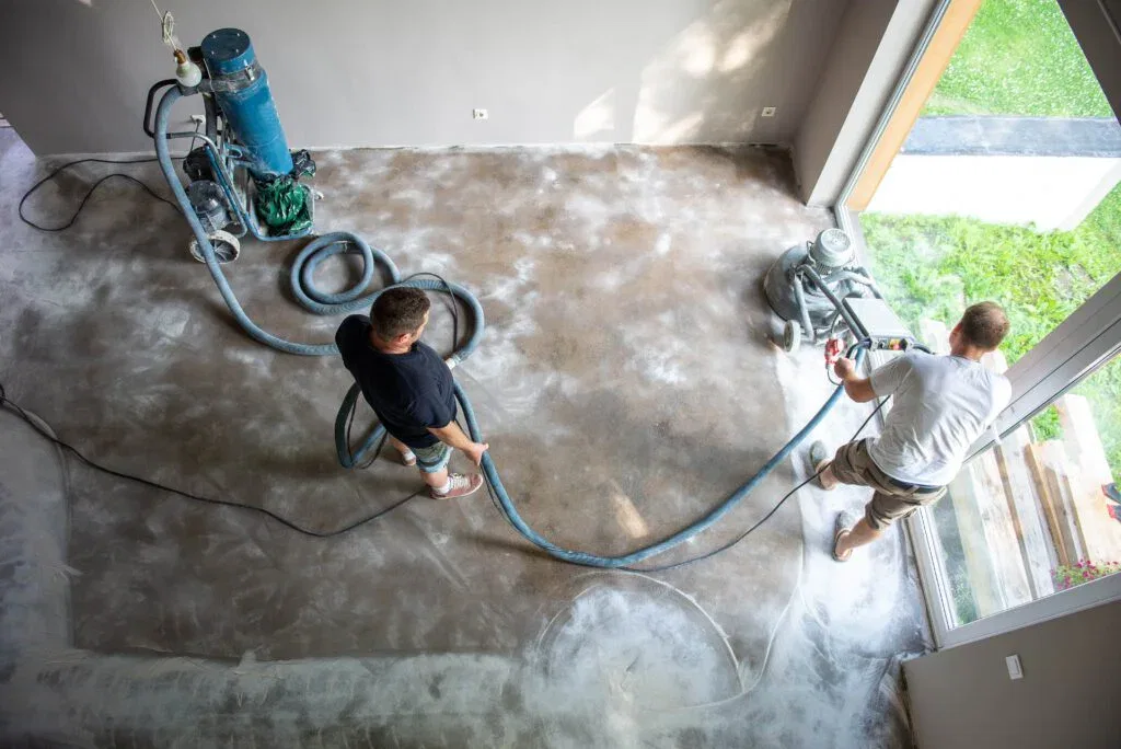Overhead view of two contractors using a professional floor grinder and a dust extraction vacuum system on a concrete floor in a residential space.
