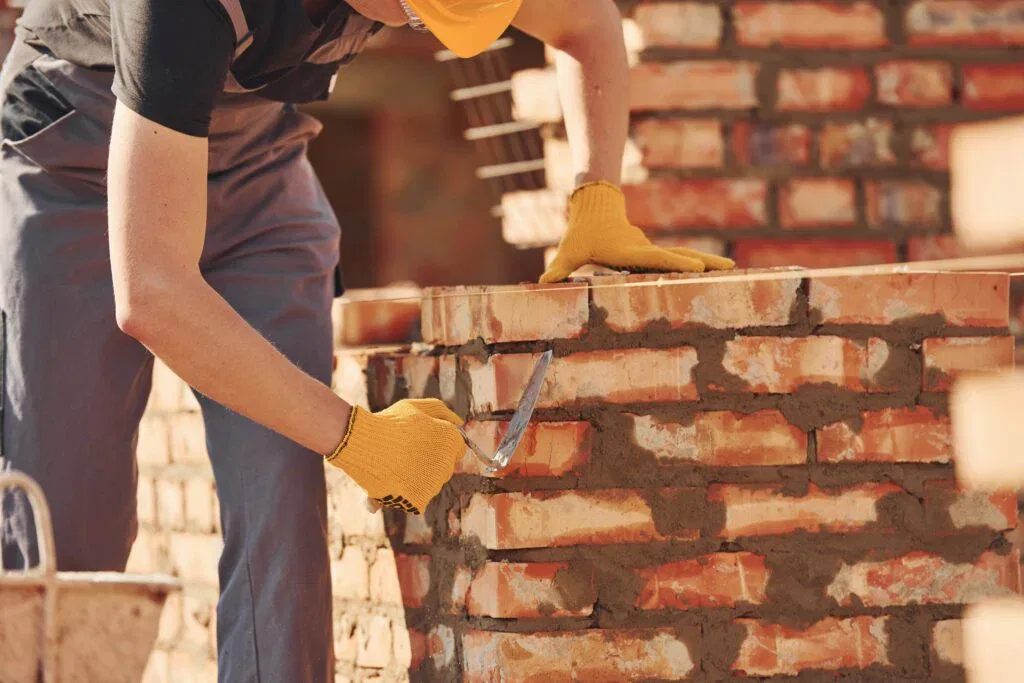 Construction worker in a yellow hard hat and safety gloves using a trowel to apply mortar while laying a red brick wall on a sunny building site.