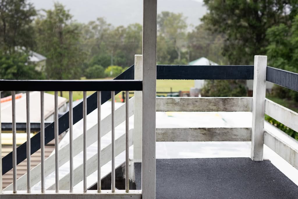 View from a wooden balcony during rain in Ocala, Florida , showing weathered white and black railings in need of maintenance with a green rural landscape in the background.