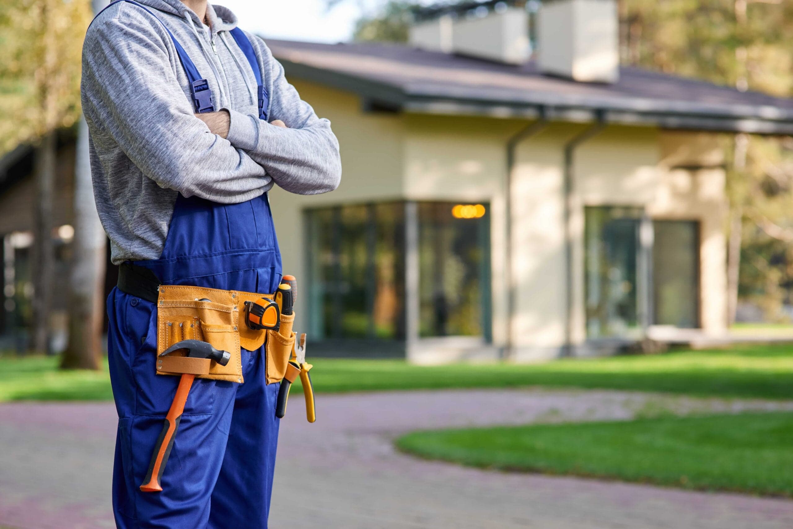 A cropped shot of a professional home renovation contractor with crossed arms wearing blue overalls and a tool belt, standing in front of a modern suburban house in Tampa, Florida.