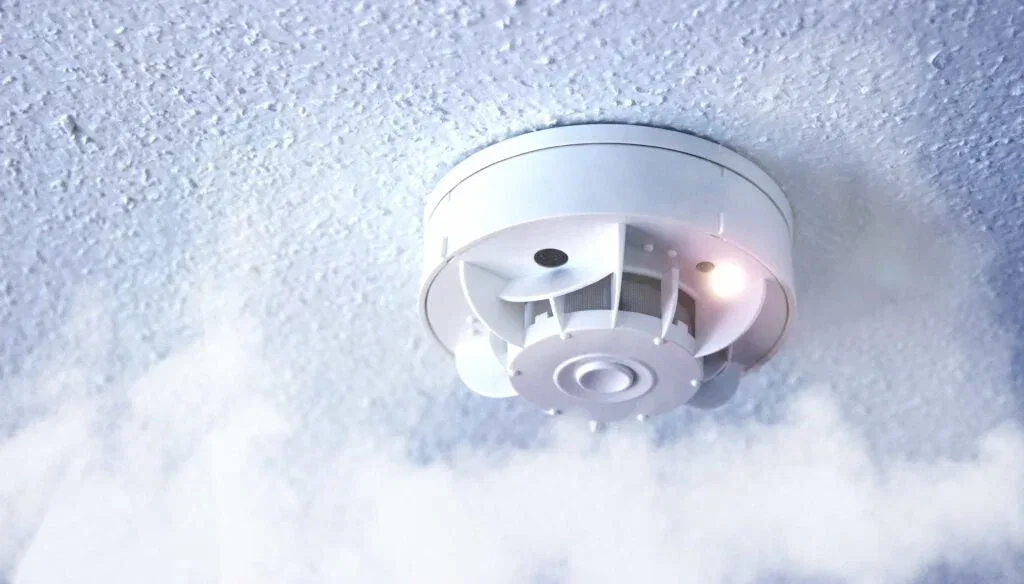 A white smoke detector mounted on a textured ceiling with visible smoke and an active indicator light. Professional residential fire safety service in Tampa, FL.